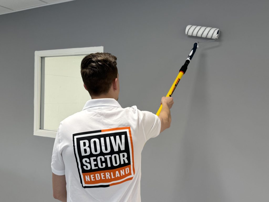 Schilder met Bouwsector-logo op zijn rug rolt een grijze muur naast een wit kozijn