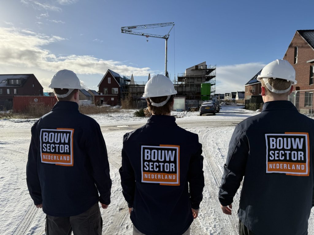 Drie vakmensen van Bouwsector Nederland in werkjas met logo bij woningen en een bouwkraan