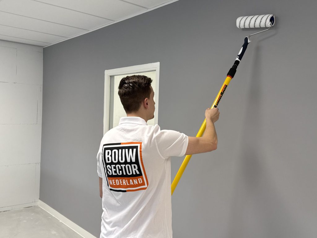 Schilder van Bouwsector Nederland rolt een grijze wand onder een plafond van platen, met links een nog kale wand